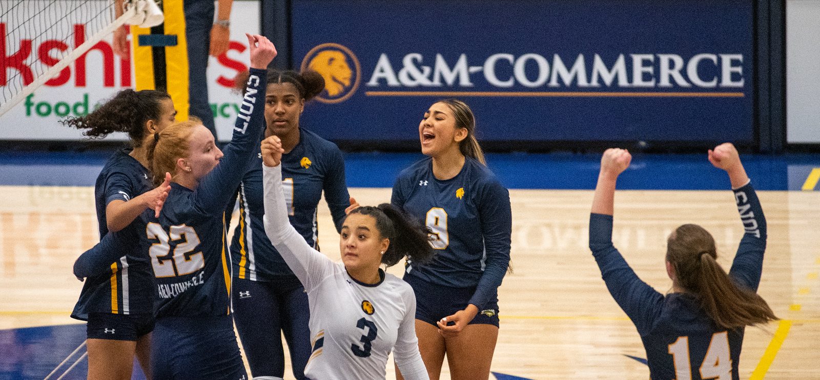 Six volleyball players cheer as they break from a huddle, with arms raised.