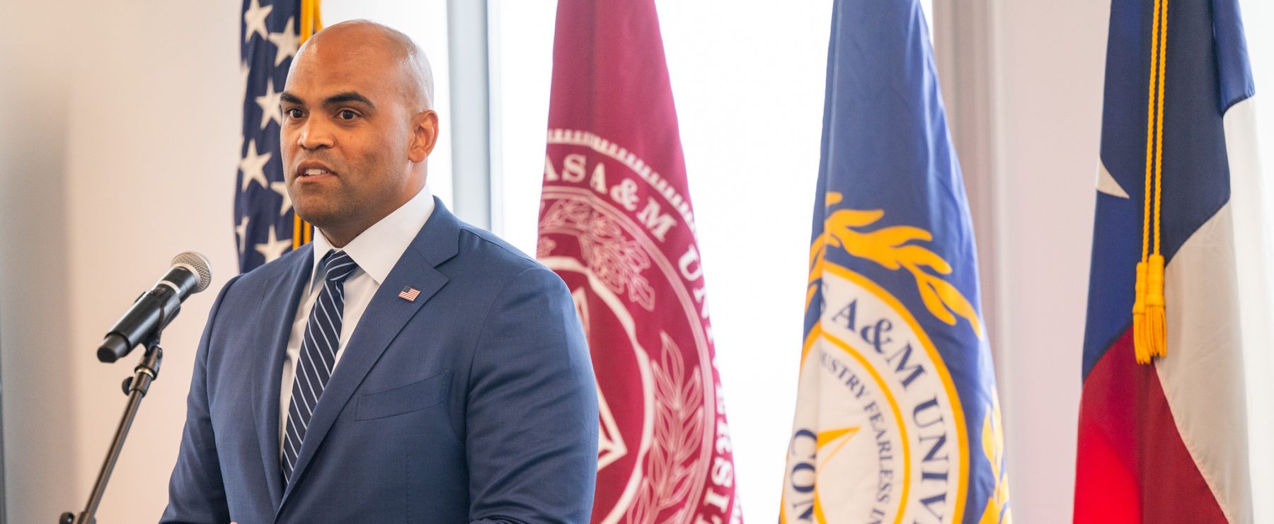 Speaker stands in front of microphone on left side of photo. Four flags are in the background.