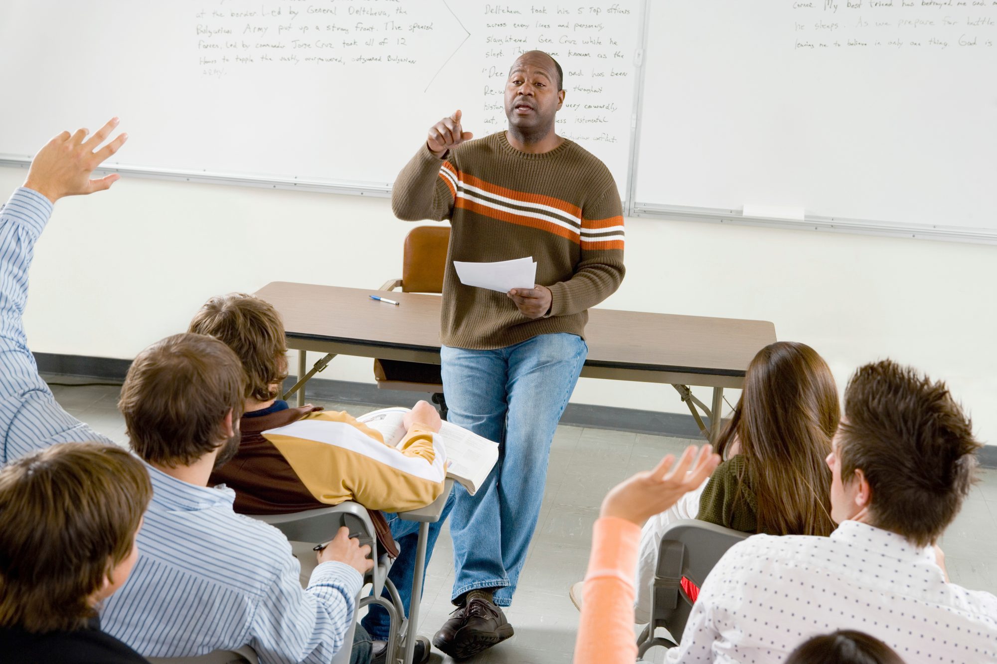 Professor pointing at college student with hands raised in classroom.