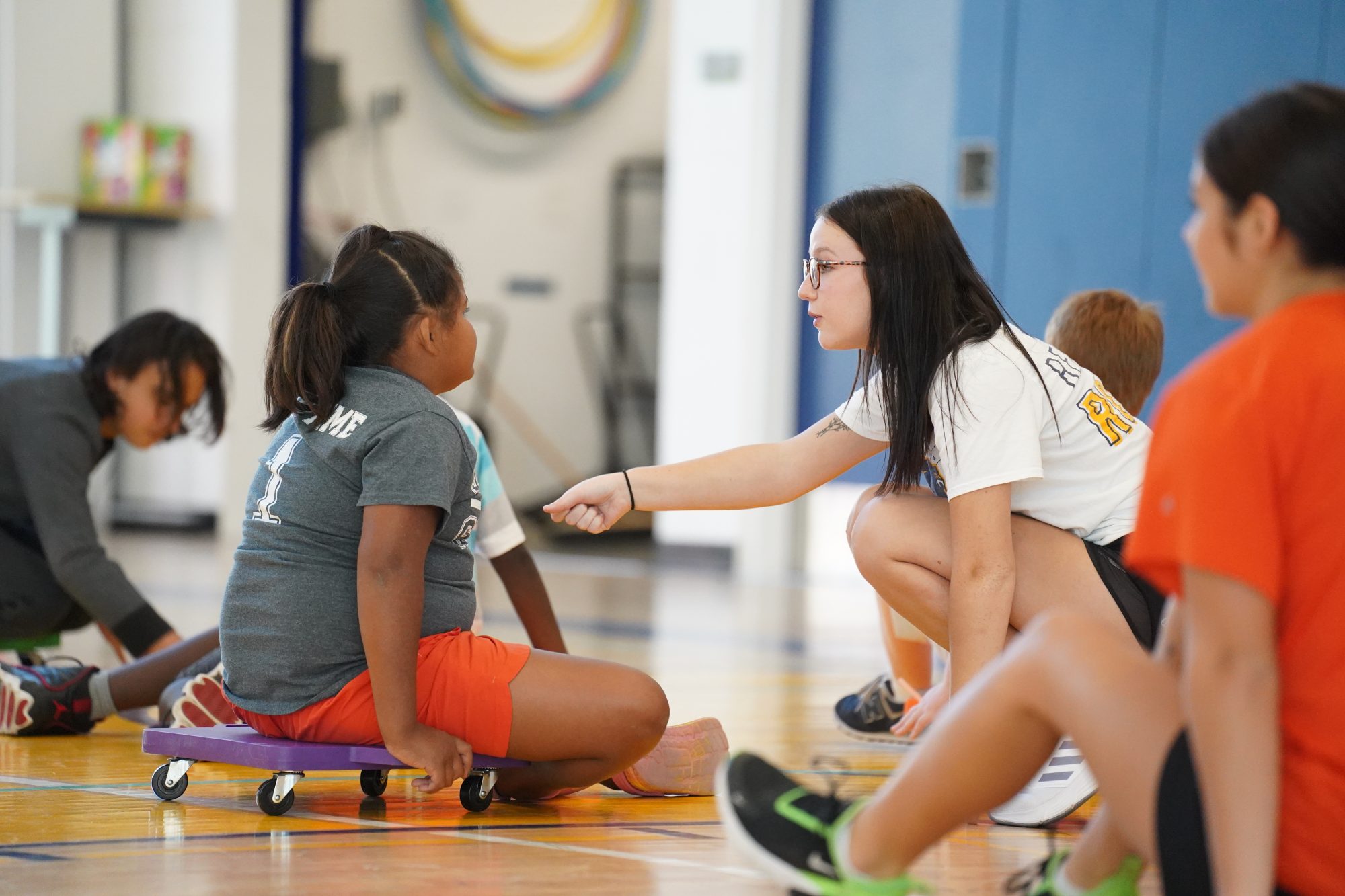 A kinesiology student provides instructions for young students participating in a gym activity on floor scooters.
