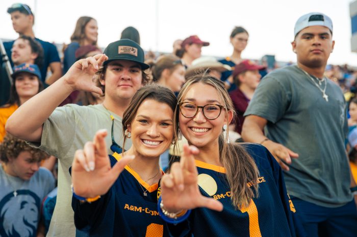 Students in bleachers at football game smile into camera and flash the Lucky sign (fingers crossed).