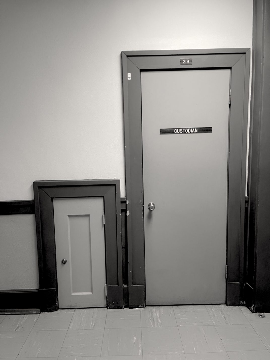 A black and white photograph of a normal door with a sign that says "Custodian," and a very tiny door with no label next to it.