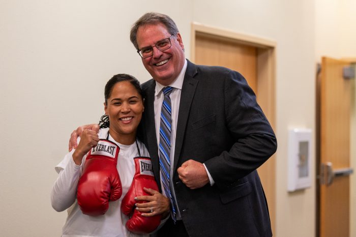 A woman with boxing gloves hanging from her shoulders poses with a man as both jokingly display fists for the camera.