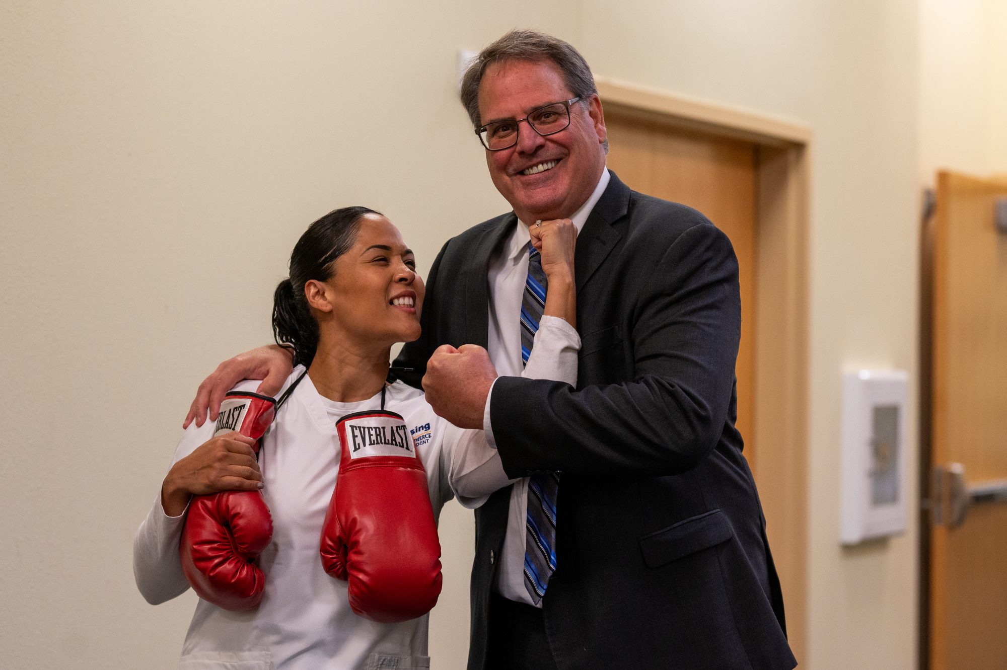 A woman with boxing gloves hanging from her shoulders poses with a man as both jokingly feign chin punches to each other.