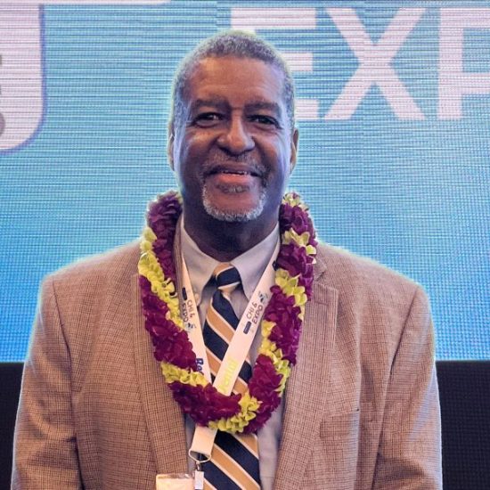 A man wearing a beige suit with a striped tie and floral lei stands in front of a blue backdrop and displays partial text.. He has short grey hair, a beard, and is smiling at the camera. A lanyard with a name badge hangs around his neck.