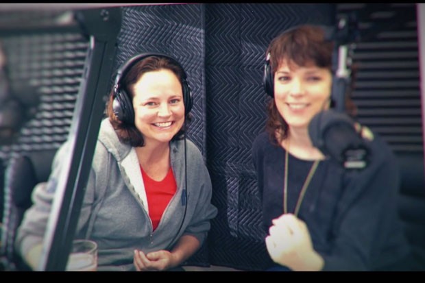 Two women sitting side by side in a podcasting booth, wearing headphones and smiling
