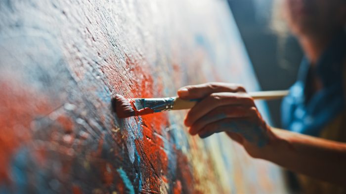 A hand holding a paintbrush and painting on a canvas