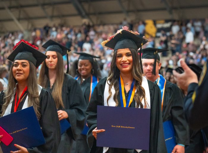 Graduates stand with their caps and gowns. Graduate in the foreground smiles with diploma in hand.