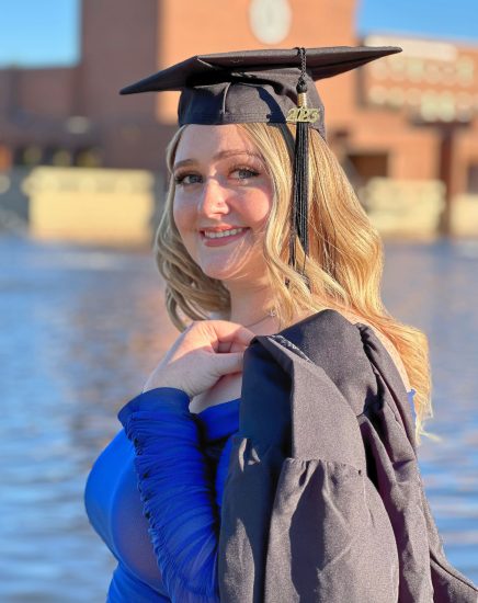 A woman wearing a commencement cap with her gown over her shoulder stands in front of Garvin Lake on the campus of Texas A&M University-Commerce.