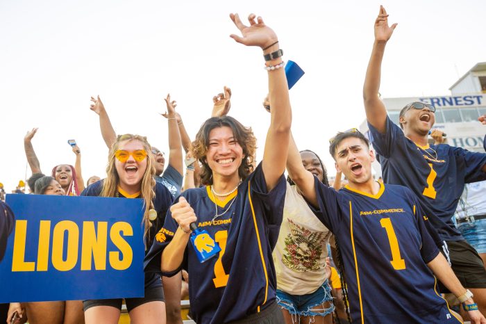 Students cheer on the football team from the stands with arms raised.