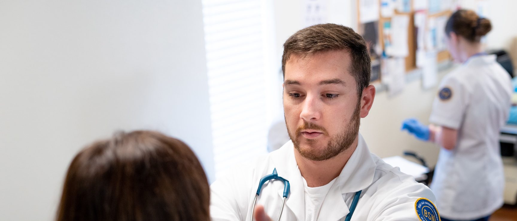 Student in white lab coat reaches out hand to patient's face during checkup.