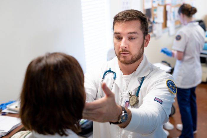 Student in white lab coat reaches out hand to patient's face during checkup.