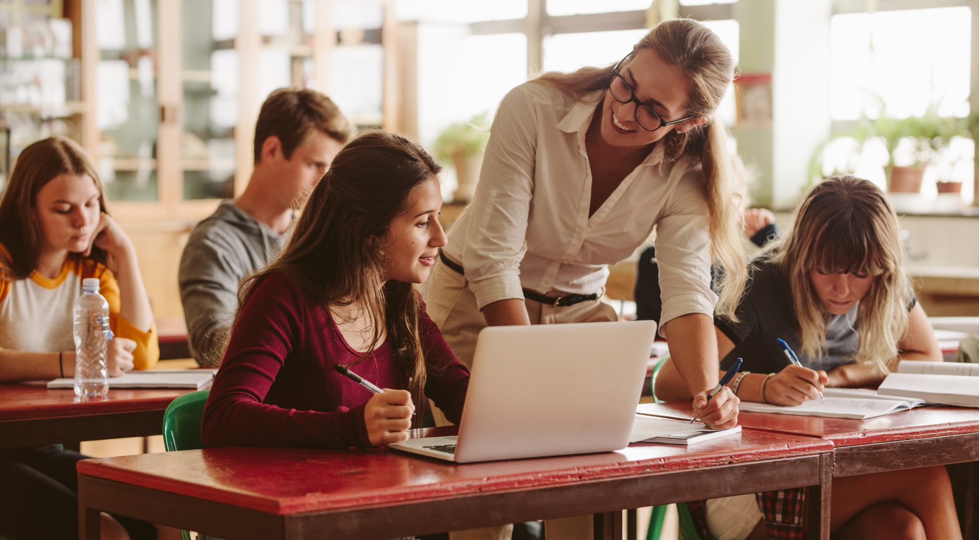 A female teacher helping a female student with a few students in the background in a classroom