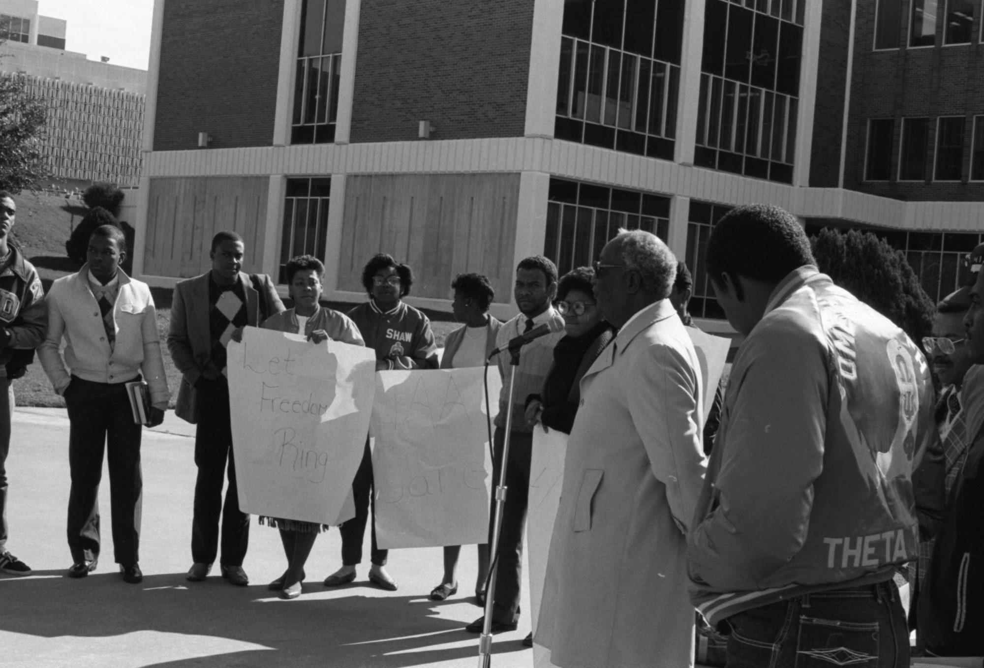 Black and white photo of David Talbot standing at a microphone with a group of all Black students holding protest signs that say "Let Freedom Ring"