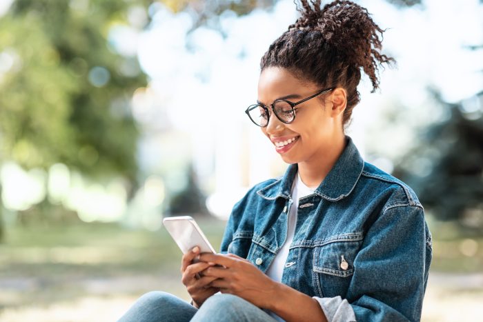 Cheerful Millennial Girl With Smartphone Using Mobile App Sitting outdoors.