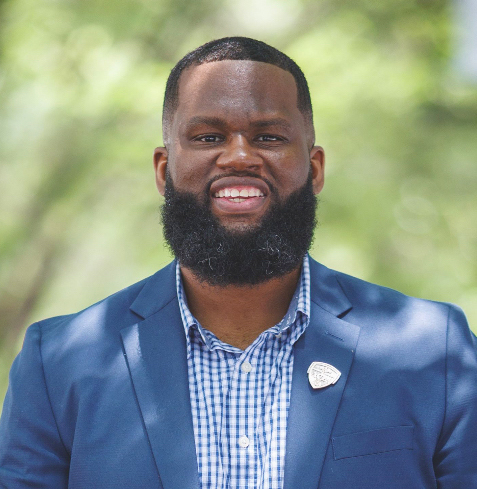 A man in a blue blazer and dress shirt stands against a backdrop of trees.