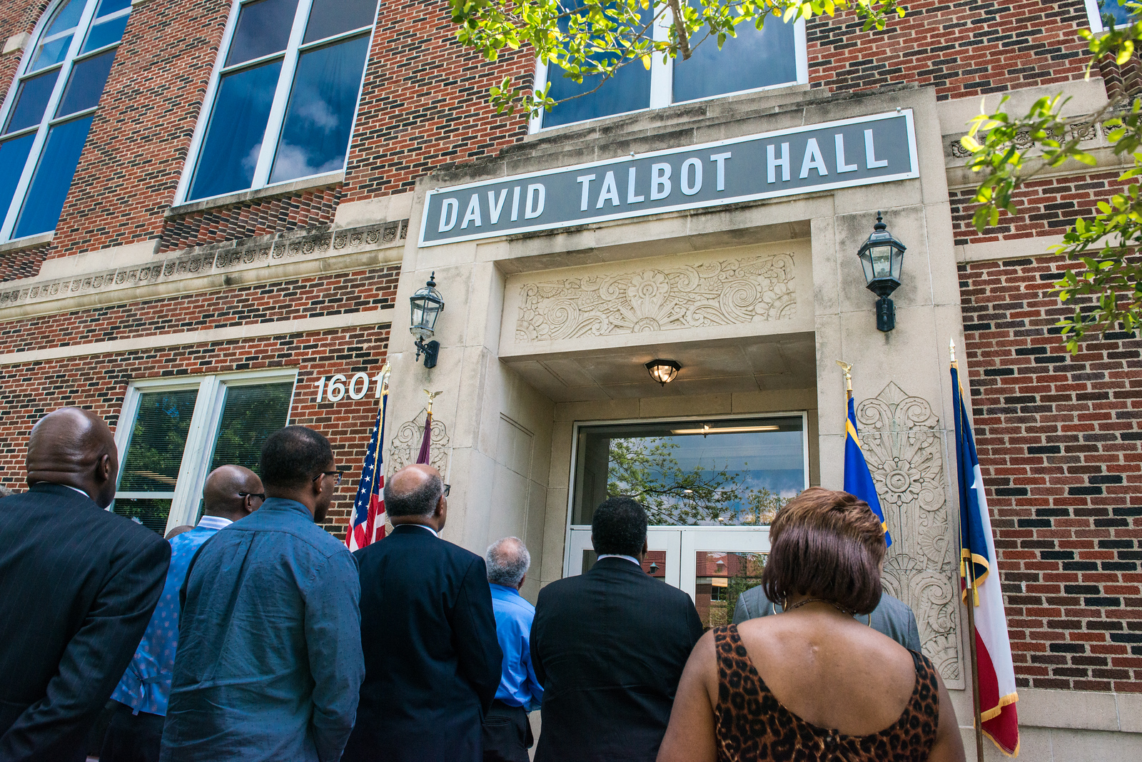 A small crowd of people looking up at a brick building with the sign "David Talbot Hall" hung above the door