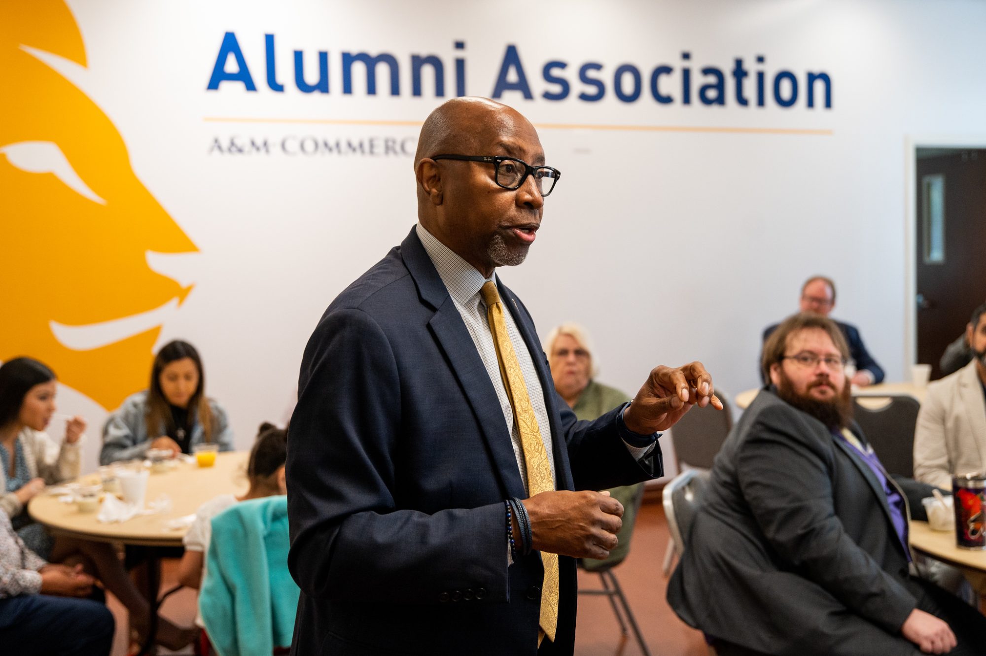 Derryle Peace presents to an audience. Audience is seated at tables all around. Audience is eating and drinking. 