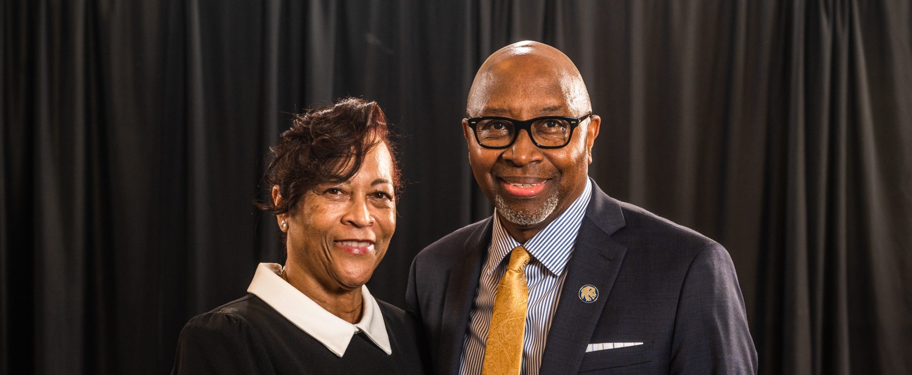 Derryle and Carolyn Peace face the camera and smile. Couple is dressed formally with black curtained background.