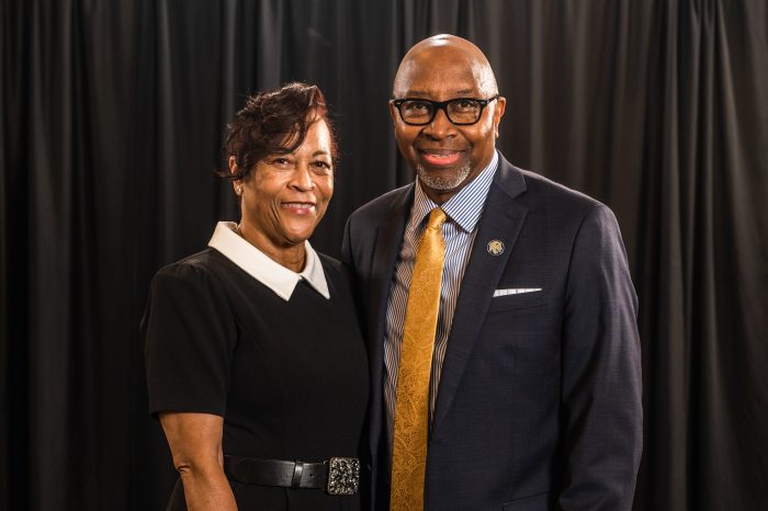 Derryle and Carolyn Peace face the camera and smile. Couple is dressed formally with black curtained background.