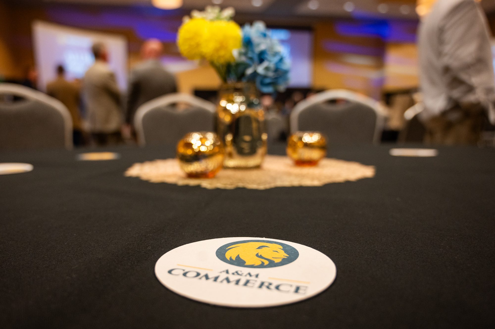A black tablecloth at the Alumni Ambassador Forum includes a gold placemat in the center with three bronze-colored ornaments on it. In the foreground, a drink coaster features the A&M-Commerce logo.