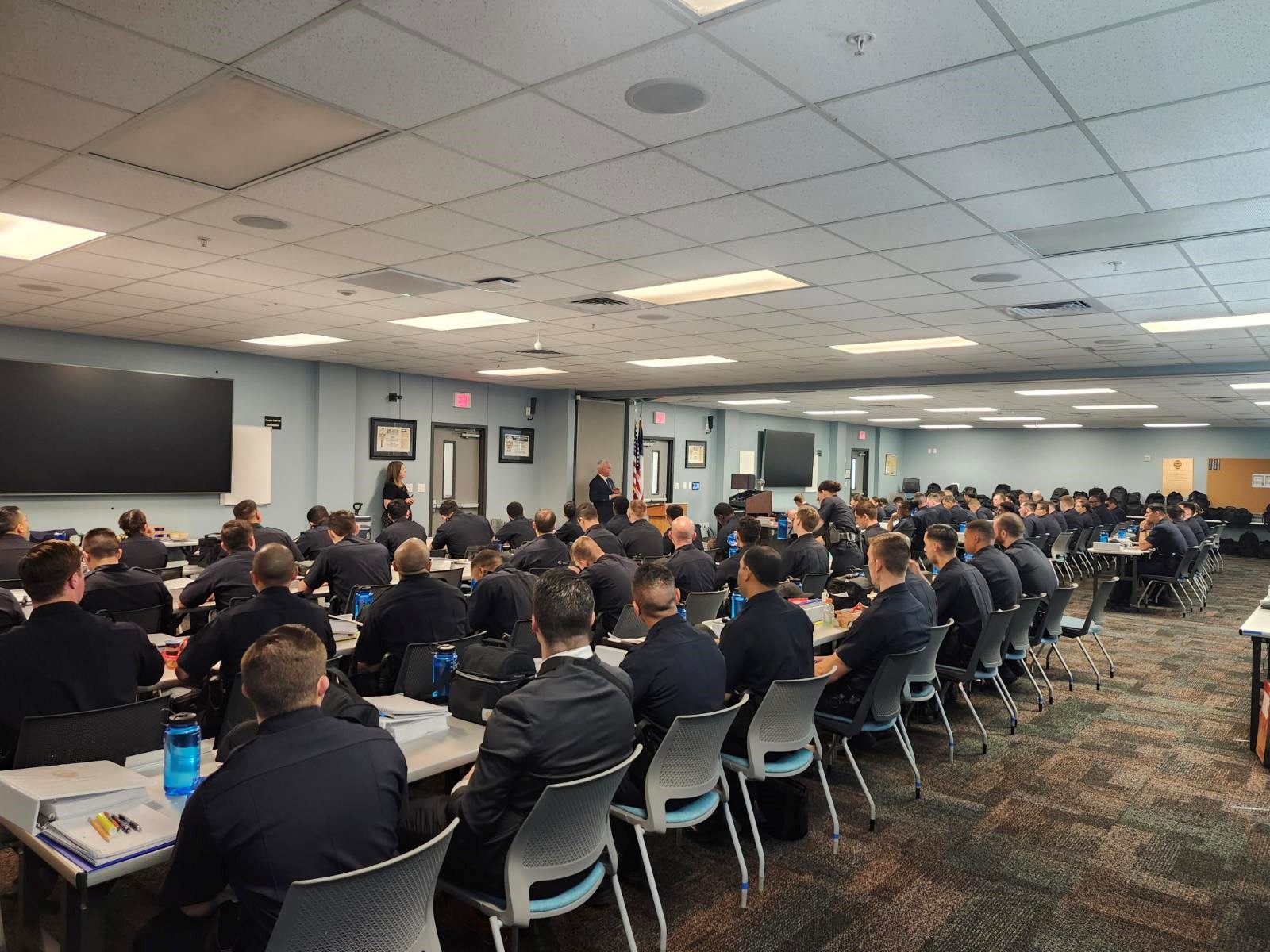 A multitude of police cadets sit at long tables listening to a presentation.