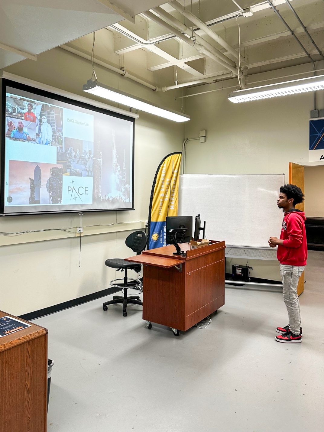 A student stands facing a projector screen displaying a video call.