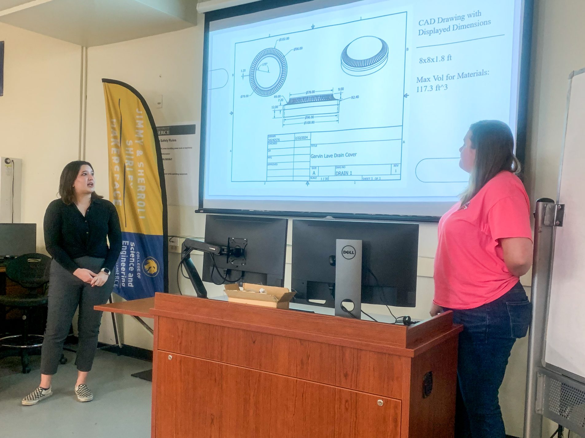 Two female students stand at a lecturn with a presentation slide showing behind them.