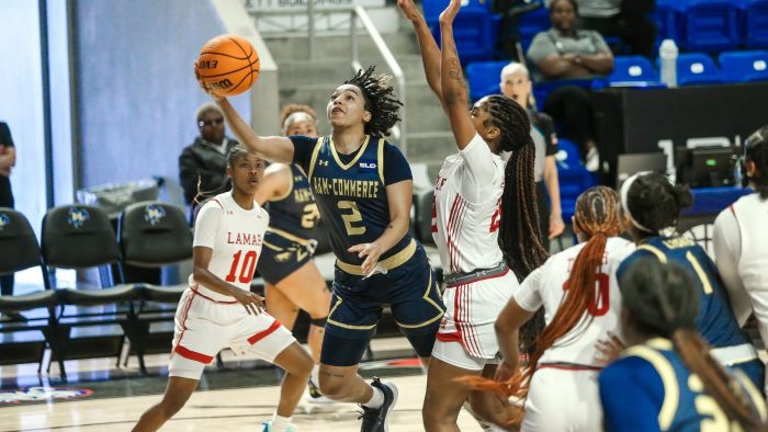 Basketball player tries to make a shot while two opponents try to block the play. Stands and spectators can be seen in the background.