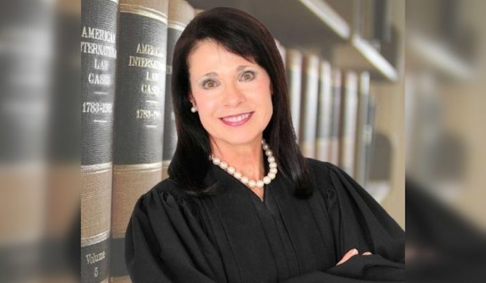 A person in a judge's robe leaning against bookshelves.