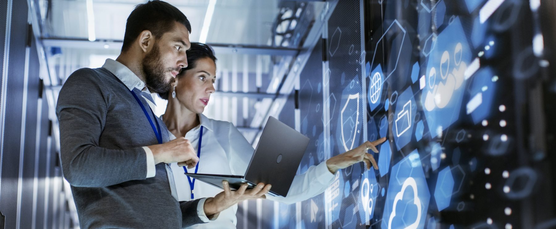 A female technology manager stands utilizing a touch screen console while her male counterpart holds a laptop at the ready.