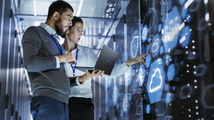 A female technology manager stands utilizing a touch screen console while her male counterpart holds a laptop at the ready.