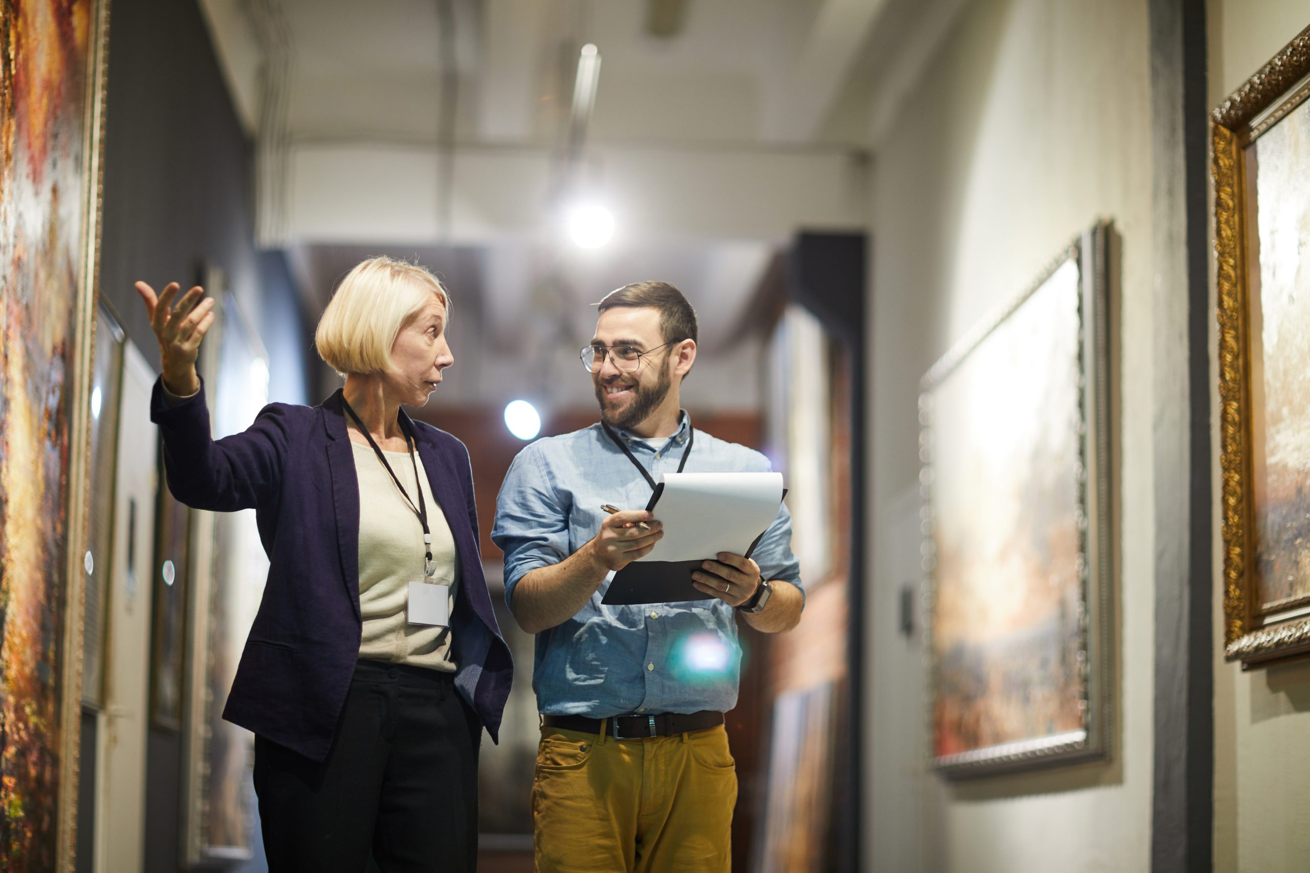 Portrait of two cheerful museum workers discussing paintings walking in art gallery, copy space.