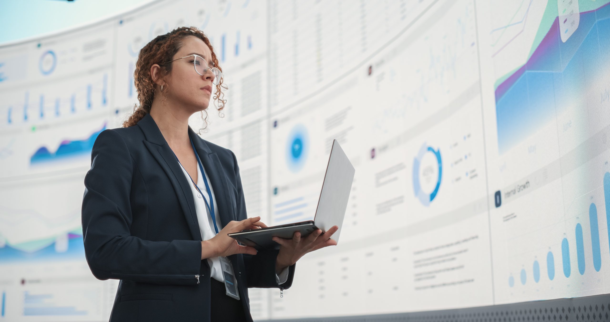 Successful Hispanic Businesswoman Holding Laptop Computer And Reviewing Financial Reports On Digital Screen In Monitoring Office. Female Leader Running Deep Learning, Artificial Intelligence Company.