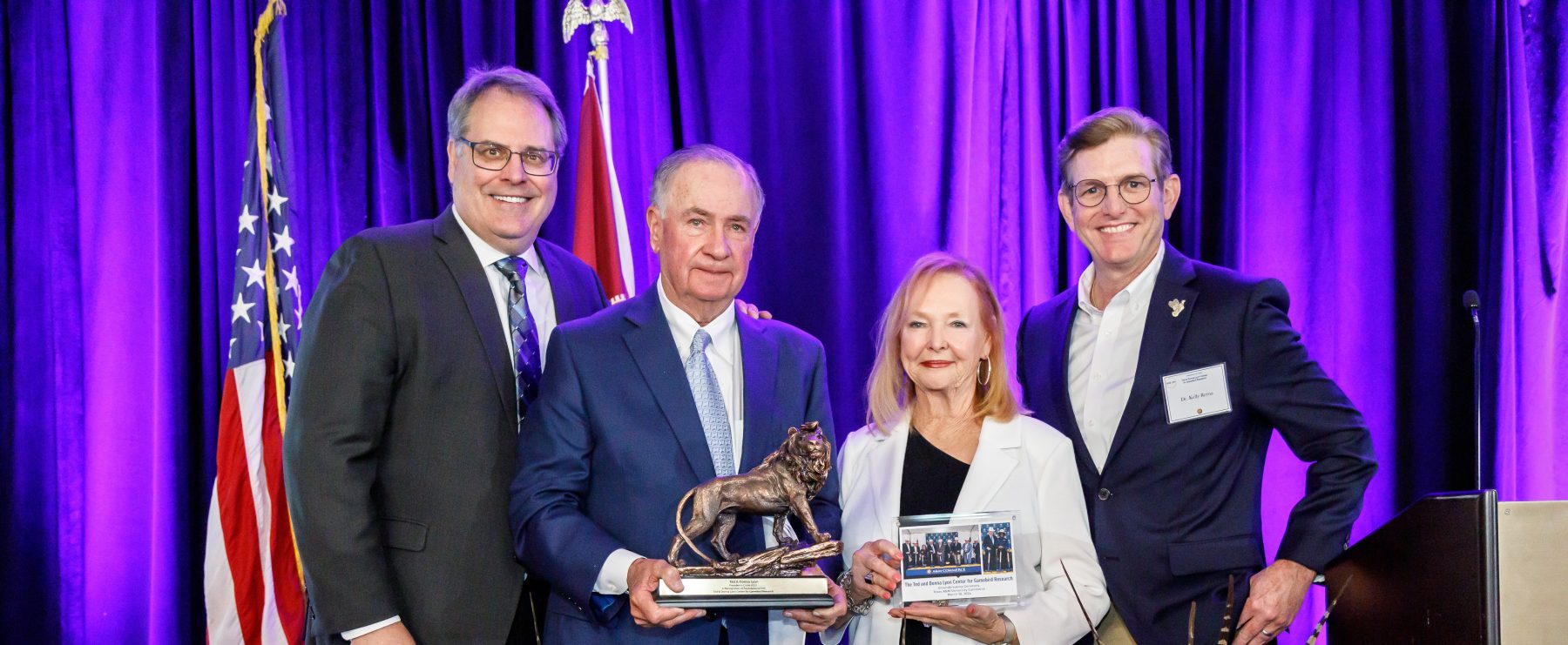 From left to right, Dr. Rudin, Senator Ted Lyon, Donna Lyon and Dr. Kelly Reyna stand on stage in front of a purple curtain and the U.S. and Texas flags. The Lyons are holding a lion statue. In front of the people, three bouquets of sunflowers sit on the floor.