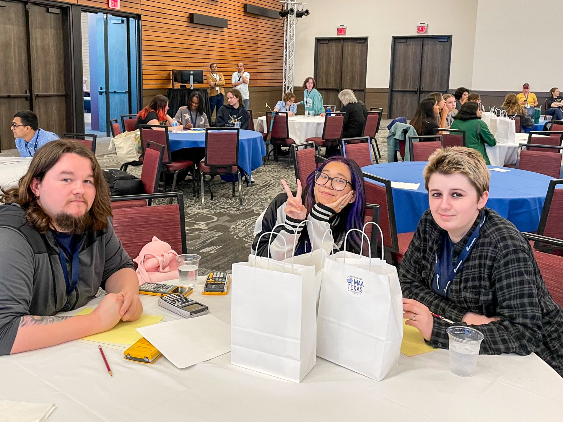 Three students sit at a table with calculators and gift bags.