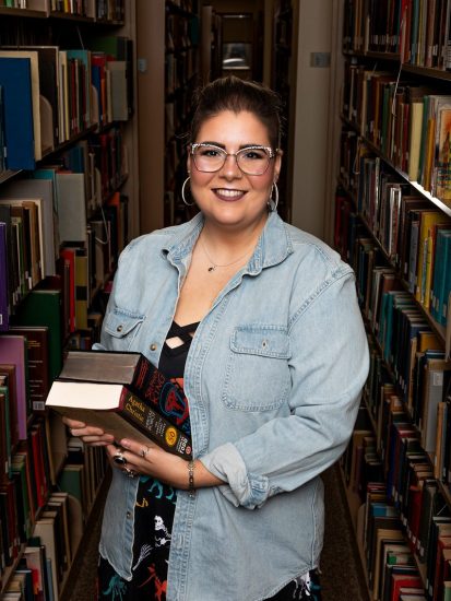 Rachel McShane standing between bookshelves holding a stack of books.
