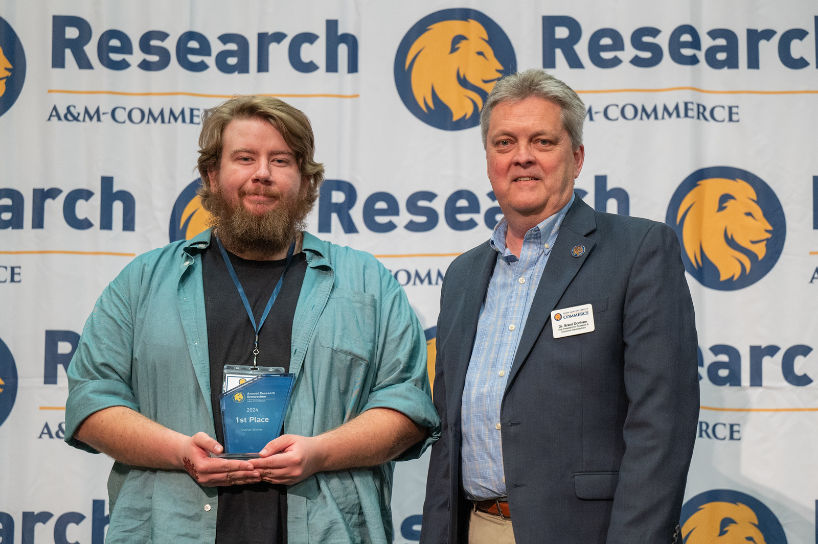 Joshua Belieu and Dr. Brent Donham stand in front of a white backdrop with A&M-Commerce Research logos, which include the university's lion head.