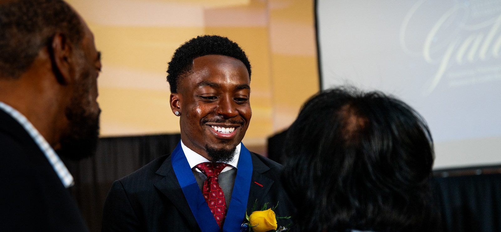 Award recipient Kader Kohou faces the camera and smiles with eyes downcast and a gold medal around his neck. Two people stand in front of him with their backs to the camera.