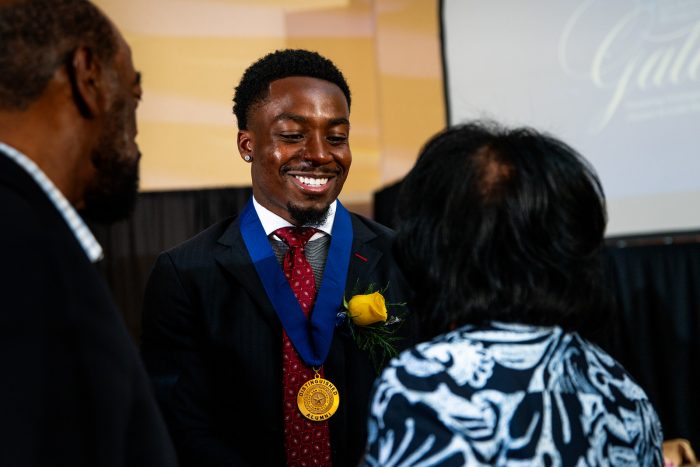 Award recipient Kader Kohou faces the camera and smiles with eyes downcast and a gold medal around his neck. Two people stand in front of him with their backs to the camera.