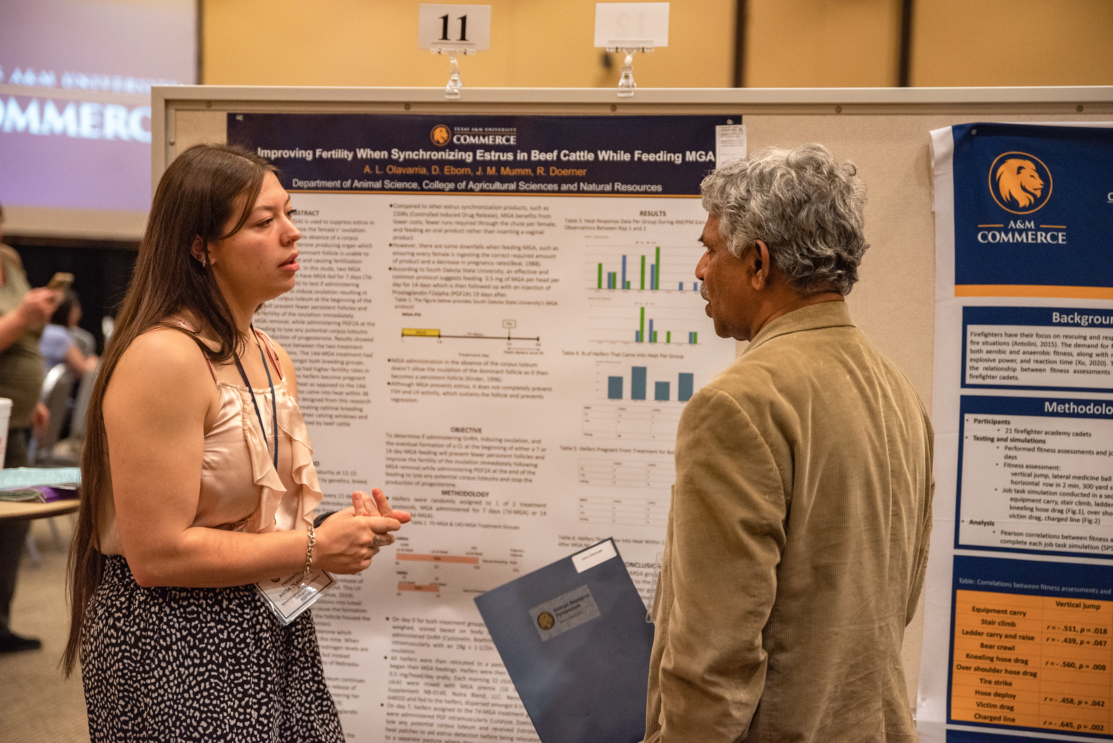 A student stands in front of her poster presentation as she converses with an individual who is holding a folder in his hand. 