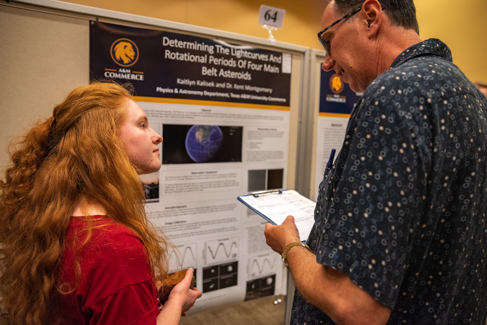 Student stands in front of her research project poster presentation and looks at a judge who holds a clipboard in his hand appears appears to ask the student a question.