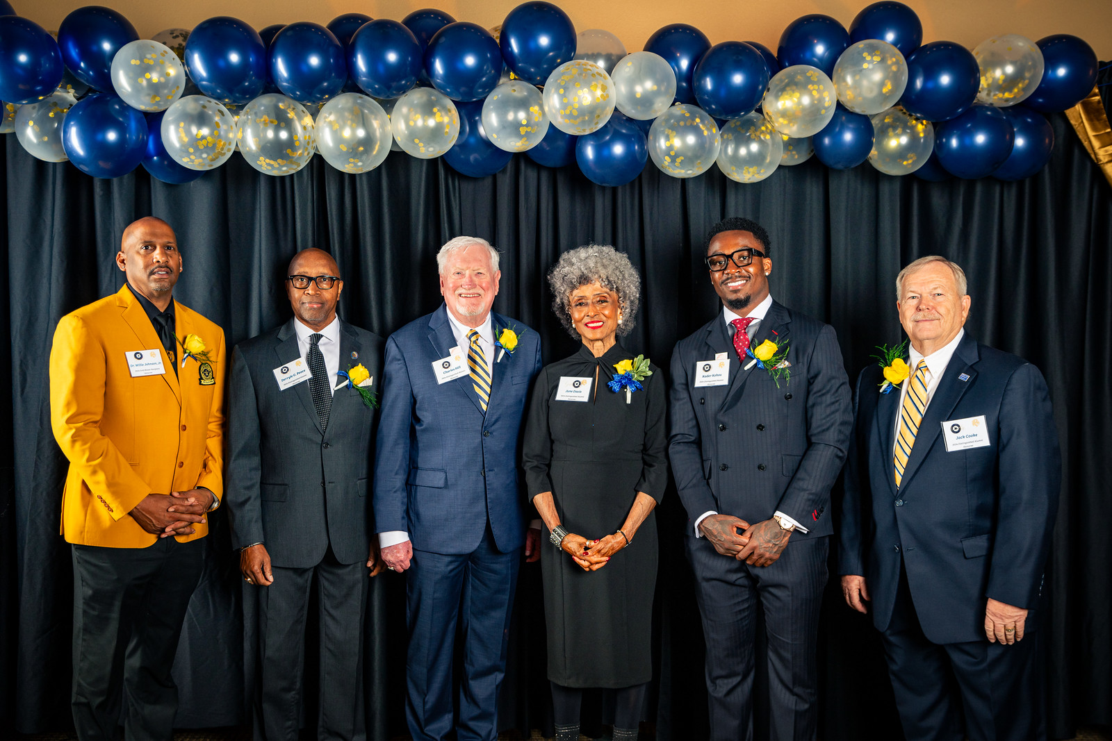 The six award recipients line up for a photo in front of a black curtain with gold and blue balloons above their heads.