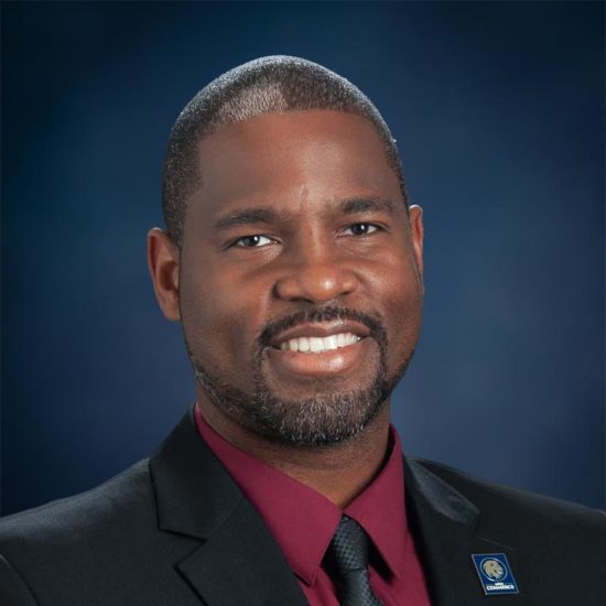 A professional headshot of a man wearing a dark suit, maroon shirt, and black tie, smiling warmly against a blue gradient background.