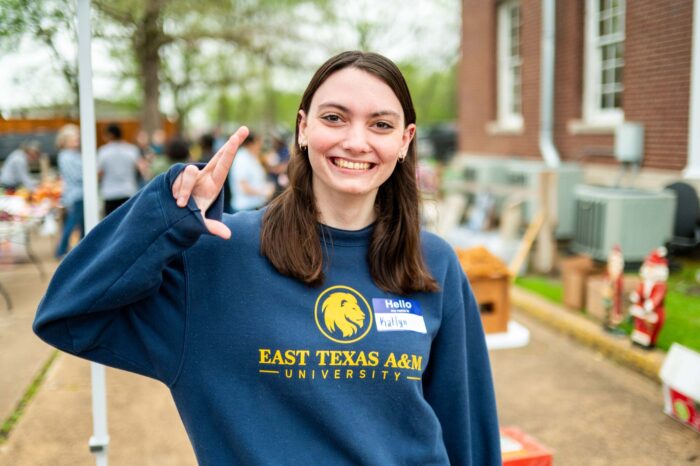 Girl smiles at the camera while making an "L" shape with her fingers. She is wearing a navy sweatshirt with "East Texas A&M" written on the front.