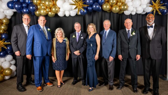 Eight individuals, dressed in formal attire, stand in front of a black curtain. Clusters of gold, blue and white ballons are above the curtains in the background.