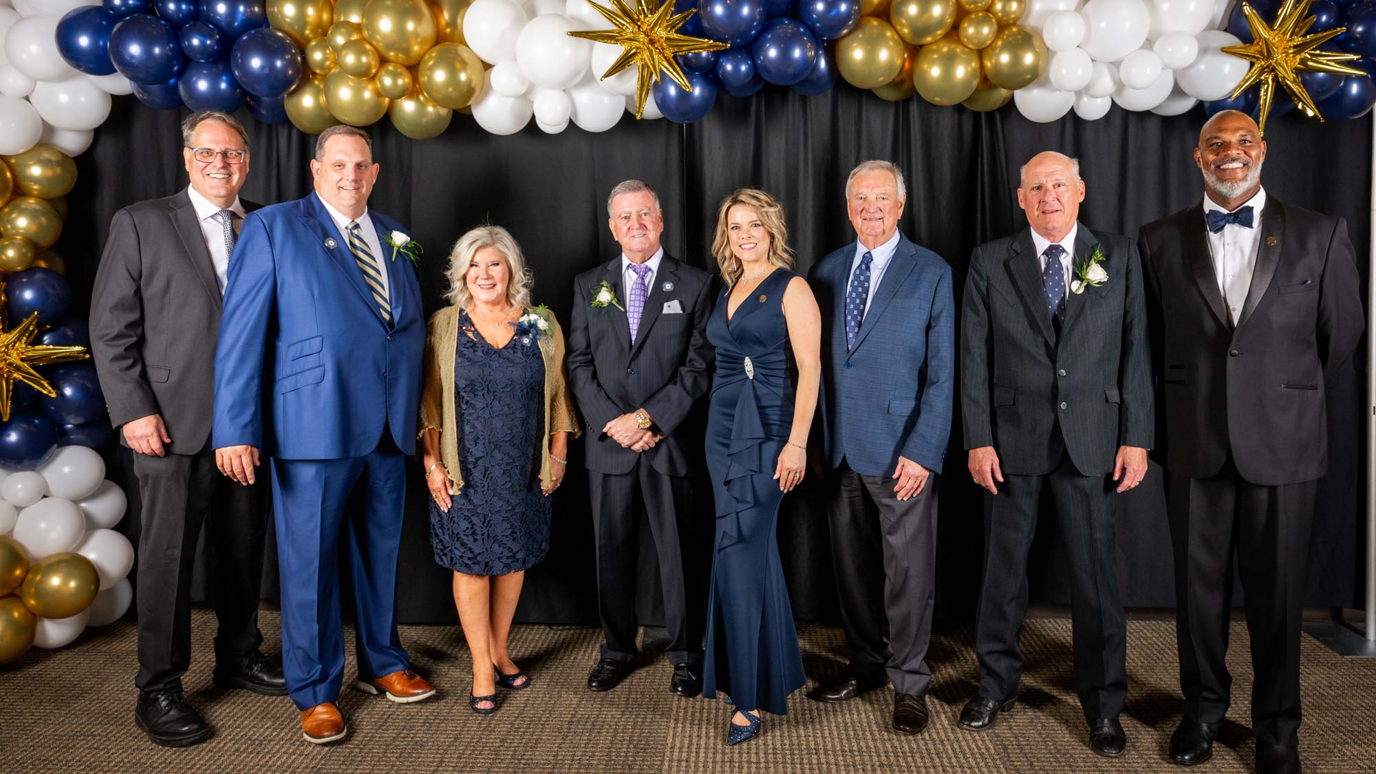 Eight individuals, dressed in formal attire, stand in front of a black curtain. Clusters of gold, blue and white ballons are above the curtains in the background.
